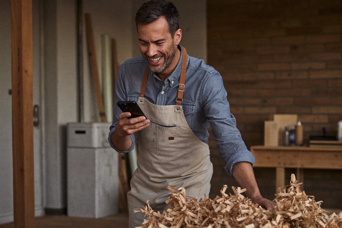 Menuisier dans son atelier, souriant en découvrant une bonne nouvelle sur son téléphone